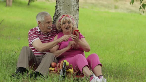 Family Weekend Picnic. Senior Old Grandparents Couple in Park Using Smartphone Online Browsing, Chat