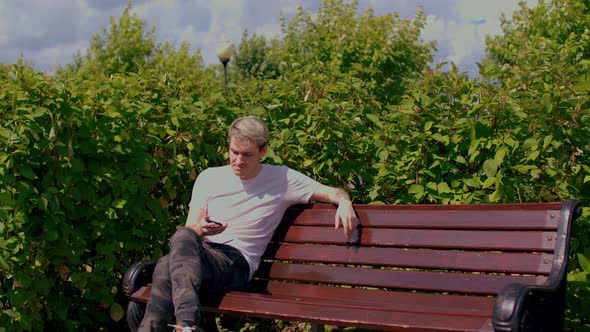 Young Man with Mobile Phone Rests Sitting on Park Bench Surrounded By Bushes