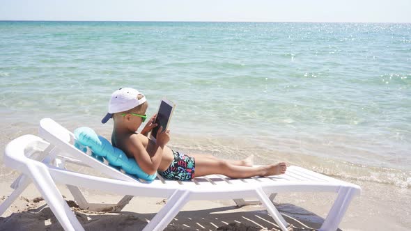 Child Trying To Play on the Tablet on a Sunny Summer Day on a Beach Chair