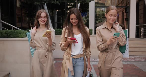 Three Cheerful Young Women Using Their Smartphones While Walking at City Street. Attractive Girls