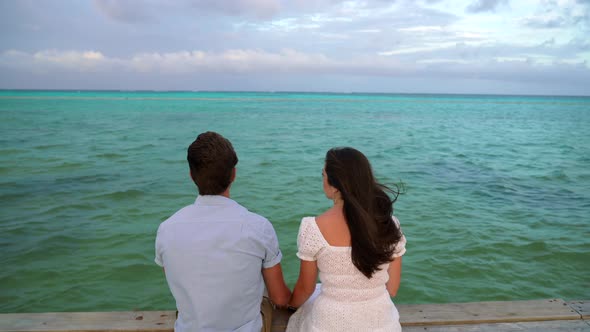 Boy and girl hold hands. Sit on wooden dock pier above turquoise tropical ocean water during sunset.