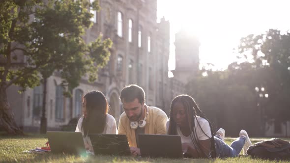 Cheerful Mates with Laptops Lying on Campus Lawn