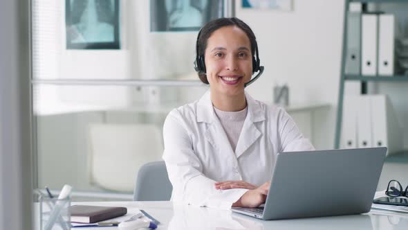 Cheerful Female Doctor in Wireless Headset Posing for Camera at Desk in Clinic