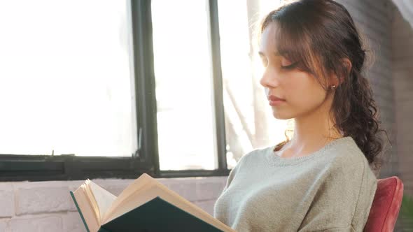 Beautiful Woman Sitting Near Window and Reading a Book on Background of Sunset