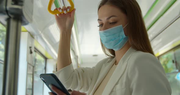 Young Woman Wearing Medical Mask To Protect Against Coronavirus Uses Her Smartphone While Traveling
