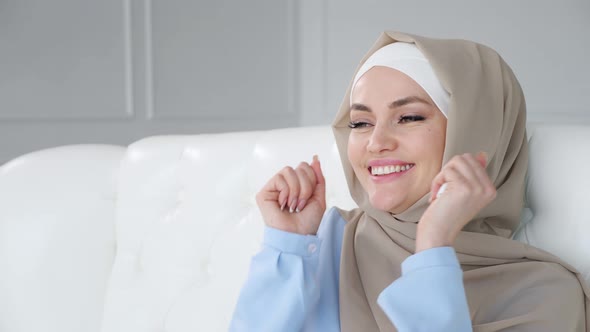 Happy Young Muslim Woman Wearing Hijab Is Dancing Sitting on Sofa and Listening Traditional Music.