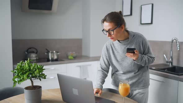 Woman with a Glass of Juice in Her Hand Stands in the Kitchen and Works at a Laptop Do Freelance