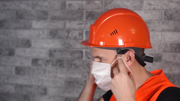 Male Construction Worker in Overalls Putting on Medical Mask on Face Background of Gray Brick Wall