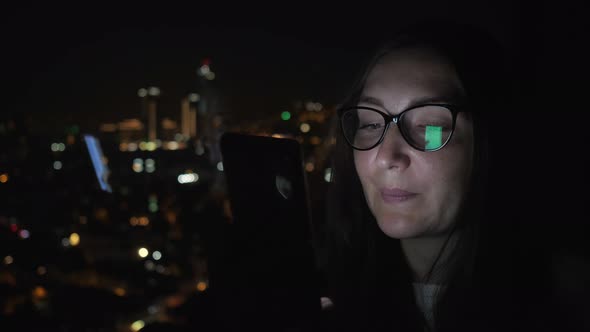 Smiling Woman is Looking at Smartphone Near the Window with City View Side View