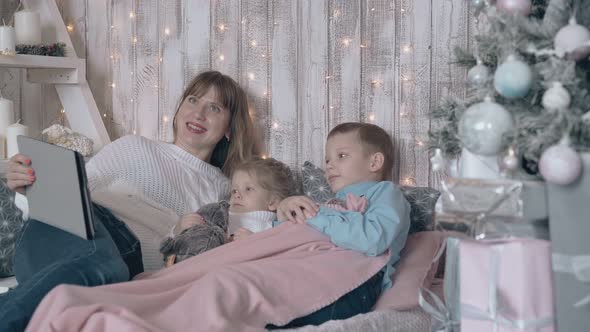 Mother with Tab and Children Lie on Sofa Near Christmas Tree