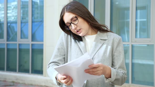 Young Woman in a Formal Suit and Glasses Talks on the Phone and Examines Documents on the Go