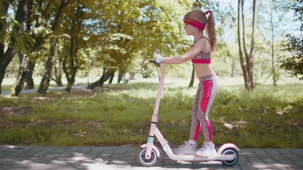 Young Athletic Fit Blonde Girl Rides Traveling on Electric Scooter on Road in Park on Sunny Day