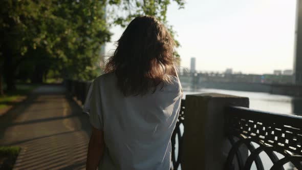 a Brunette in Casual Clothes Goes and Touches Hair in a City Park Near an Openwork Lattice