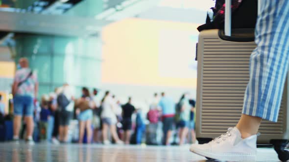 Close-up of Walking Legs in Sneakers, with Suitcase on Wheels, Through Airport Lobby, Against