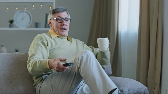 Greyhaired Aged Man in Glasses Sitting on Couch Watching TV Enjoying Relaxing at Home Drinking Tea