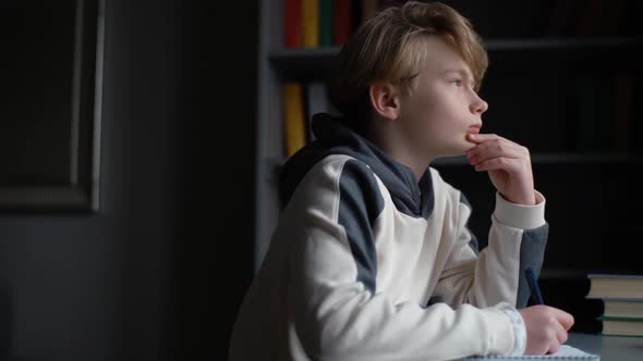 Closeup View of Thoughtful Smart Schoolboy Making Notes Writing Essay in Notebook Sitting at Desk