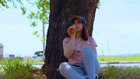 Young Woman Rests and Drinks Apple Juice Sitting Near Tree on Street