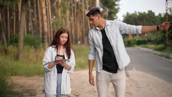 Handsome Man Gesturing Hitchhiking As Cheerful Woman Writing Destination on Cardboard Banner