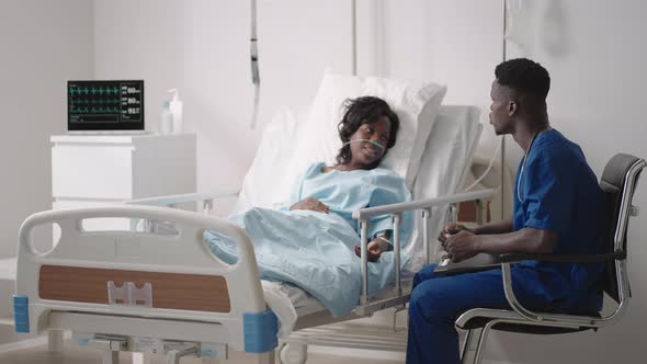A Black Male Doctor is Talking to a Black Female Patient Lying on a Hospital Bed and Connected to an