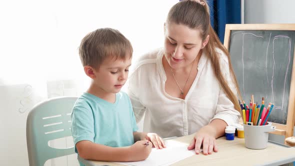 Smiling Mother Looking at Her Little Son Drawing or Writing While Sitting Behind Desk at Home