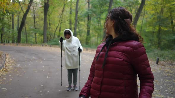 Smiling Mature Charming Woman Looking Back at Friend Endorsing Sportswoman and Strolling with Poles