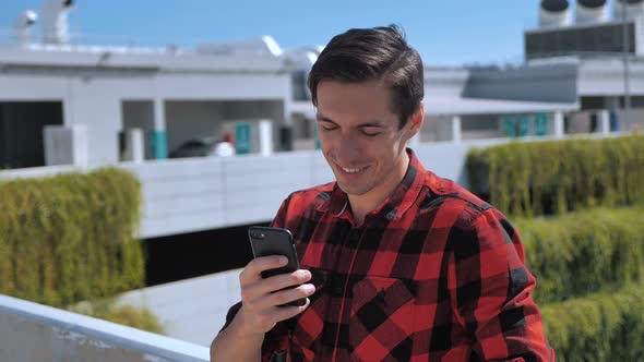 Young Smiling Man in Plaid Shirt Texting Message on Telephone Modern Smart Phone at Roof on Urban