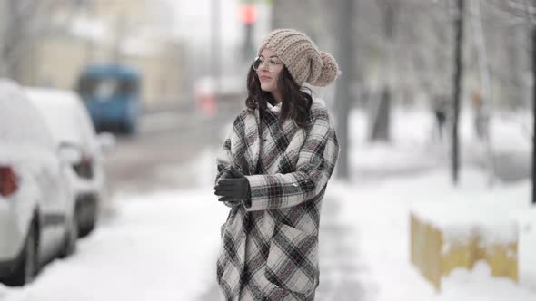 A Beautiful Young Darkhaired Woman in Round Glasses and a Plaid Coat Walks Around the City on a