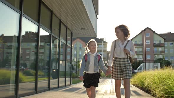 Two Schoolgirls of Different Ages with Backpacks Walk From School After School Holding Hands