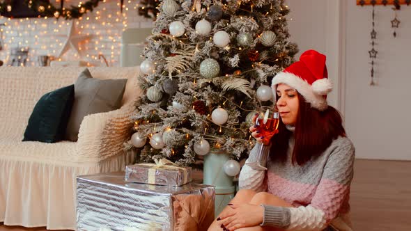 Young Woman in Santa Hat Sitting on Floor Surrounded By Gift Boxes and Christmas Tree