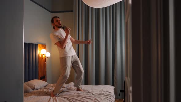 Bearded Guy with Long Hair Has Fun Dancing and Singing on the Bed in the Bedroom in the Evening