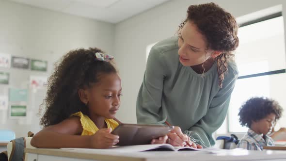 Video of happy caucasian female teacher explaining lesson to african american girl holding tablet