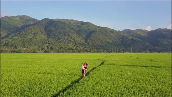 Aerial View Woman Carries Girl Among Fields Against Mountains