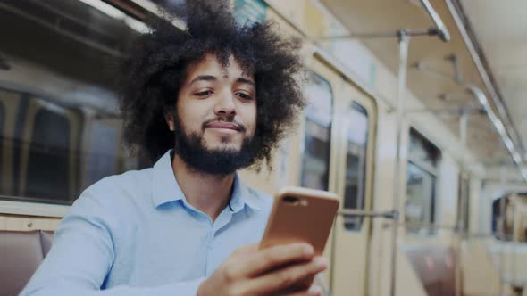 Stylish Expressive Curly Dark-Skinned Man Sitting in Subway Wagon with Smartphone. Riding at Public