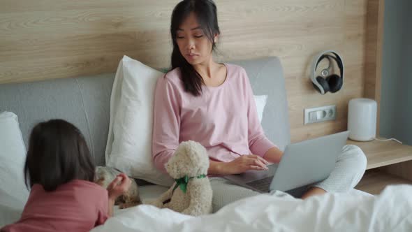 Concentrated Asian mom working on laptop while her daughter stroking dog