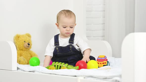 Baby Boy in Denim Overalls Sitting on the Bed Plays Toys and Throws Plastic Colored Balls