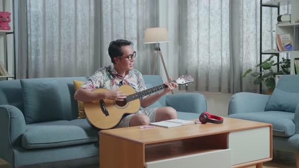 Asian Man Composer With Notebook On Table Singing And Playing Guitar At Home