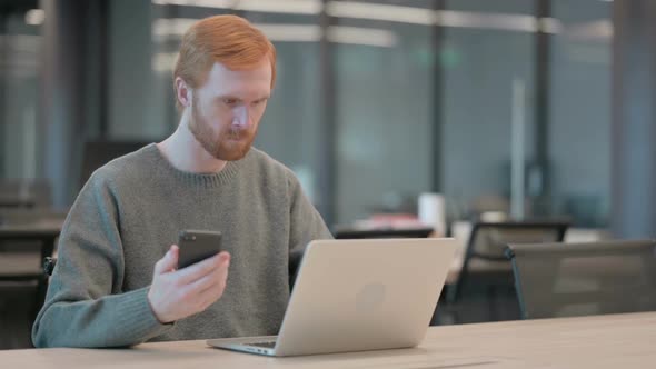 Young Man Using Smartphone While Using Laptop in Office