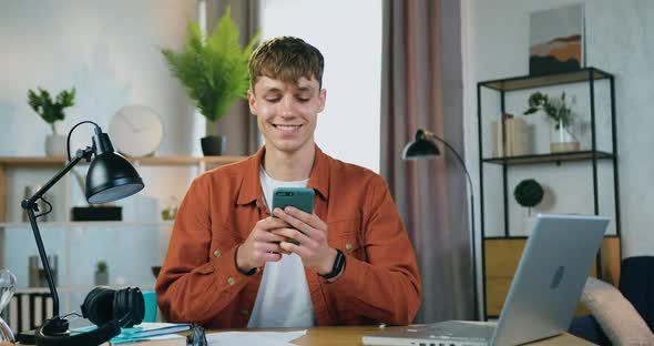 Young Guy Sitting at the Table at Home and Uses His Mobile to write Message to His Mates