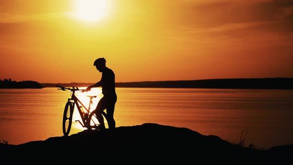 Professional male biker gets off the bike near the river at a beautiful sunset.