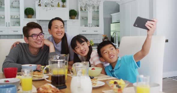 Happy asian parents in kitchen having breakfast with son and daughter, son taking group selfie