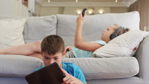 Portrait of caucasian brother and sister lying on sofa using tablet and smartphone