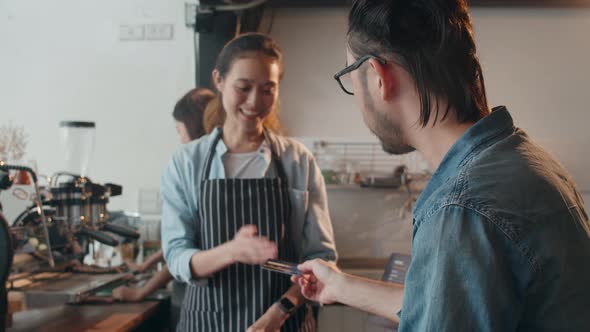 Young Asia female barista serving take away hot coffee paper cup to consumer and using credit card