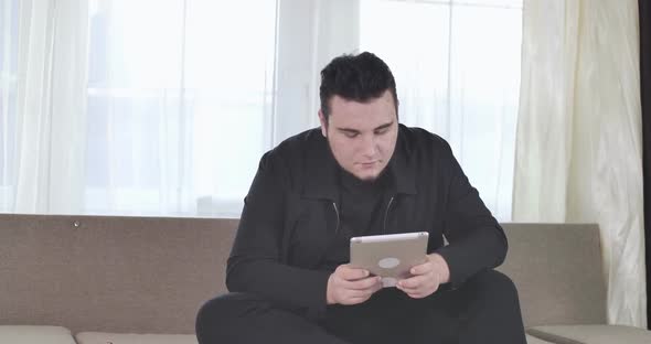 Portrait of Plump Brunette Caucasian Man Sitting on Couch Indoors and Using Tablet