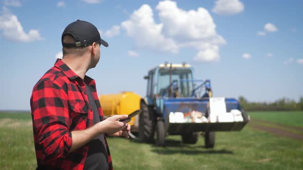 Young Attractive Farmer with Phone Standing in Field, Tractor Working in Green Field in Background