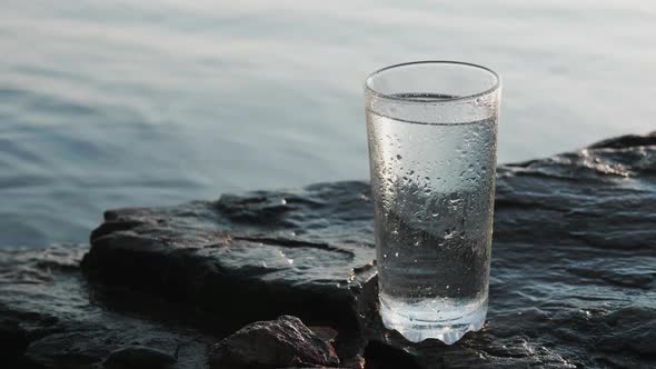 Drink in Transparent Tall Glass Stands on Stony Seashore Under the Bright Sun