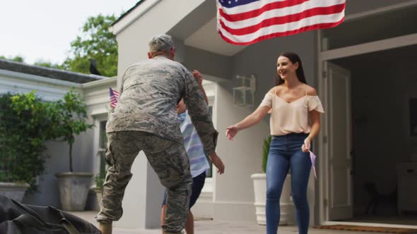 Happy caucasian male soldier lifting son and greeting wife with flag hanging outside their house