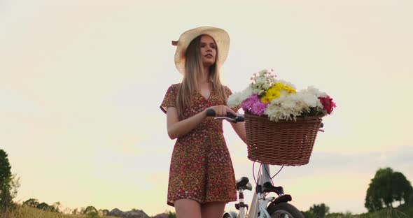 Porter Girl Walking on Camera in a Dress with Flowers in a Basket and a Bike in the Field