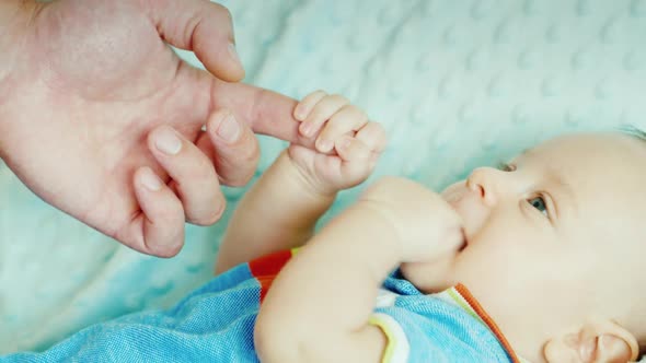 Carefree Baby Two Months Gnawing His Fist, Holding the Finger of His Father's Hand. Lying in Bed