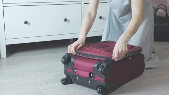 Unrecognizable Female Hands Zipping Red Suitcase on Grey Floor of Room Indoors