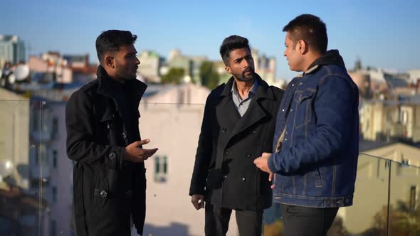 Three Positive Male Friends Talking Standing at Glass Fence on Rooftop